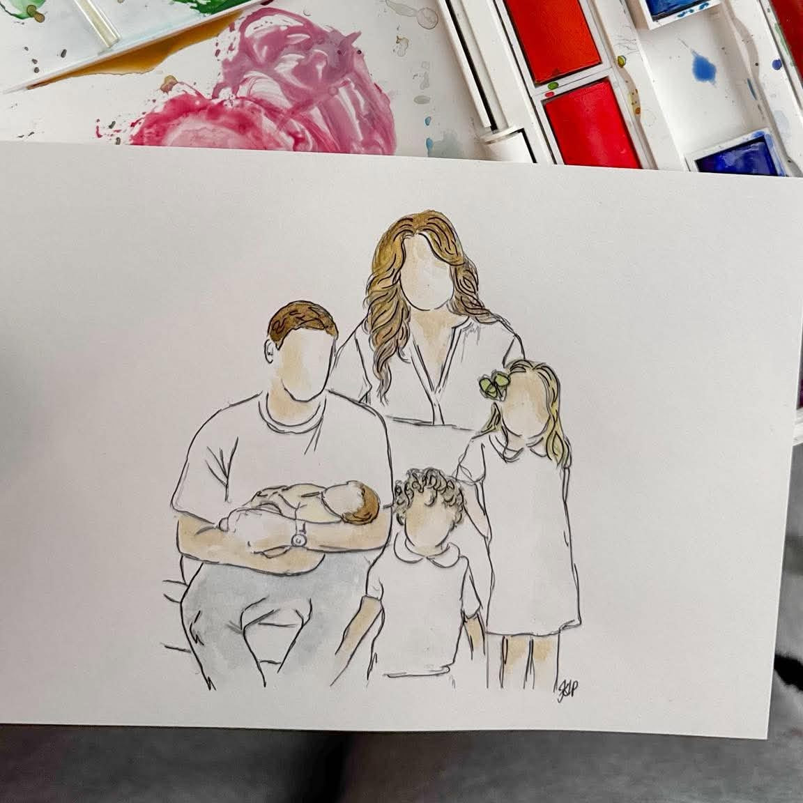 A watercolor painting of a family on a white sheet of paper, shown in front of an open watercolor palette with mixed paint colors.